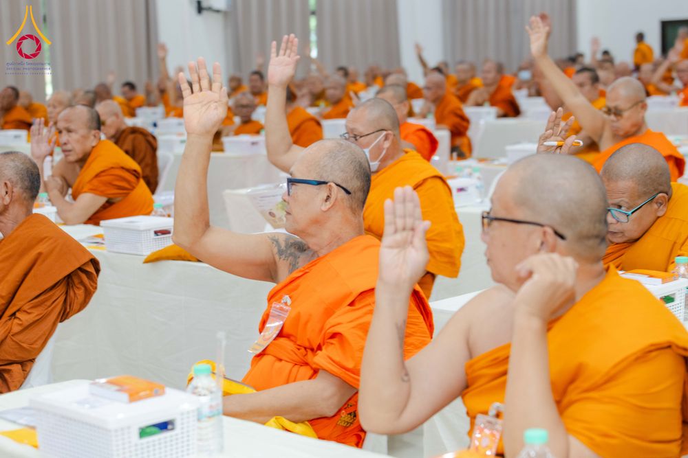 พระภิกษุสงฆ์หลายรูปนั่งเรียงกันในห้องประชุมและยกมือขึ้นพร้อมกัน