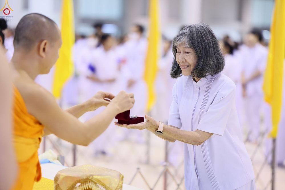 หญิงสูงวัยสวมชุดสีขาวกำลังรับมอบกล่องพระของขวัญจากพระภิกษุสงฆ์ในพิธีทางศาสนา
