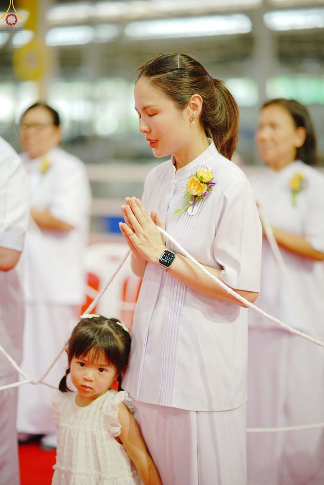 หญิงสาวพนมมือและเด็กหญิงยืนอยู่ข้างกันในงานพิธีที่มีผู้คนสวมชุดสีขาว