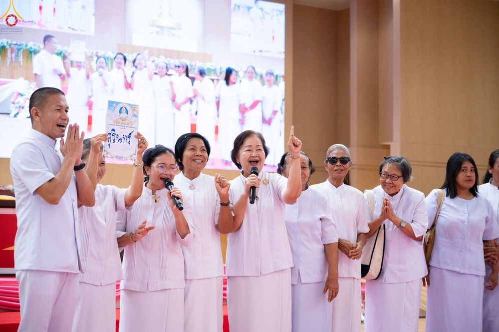 กลุ่มคนในชุดสีขาวกำลังทำกิจกรรมร่วมกันในงานสัมมนา มีผู้หญิงถือป้ายและพูดผ่านไมโครโฟนด้วยสีหน้ายิ้มแย้ม