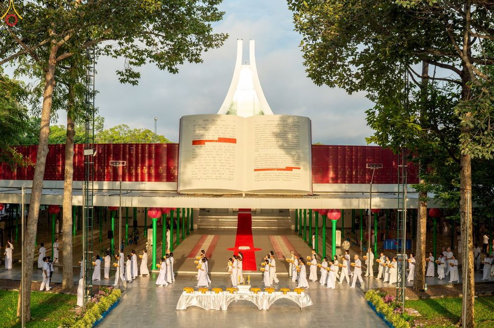 กลุ่มคนในชุดขาวเดินแถวเข้าสู่โบสถ์พระไตรปิฎกที่มีสถาปัตยกรรมรูปหนังสือขนาดใหญ่