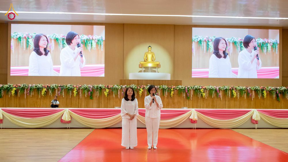 สุภาพสตรีสองท่านยืนบนเวทีในห้องประชุมที่มีฉากหลังเป็นจอภาพแสดงภาพของพวกเธอและพระพุทธรูปองค์เล็กตรงกลาง