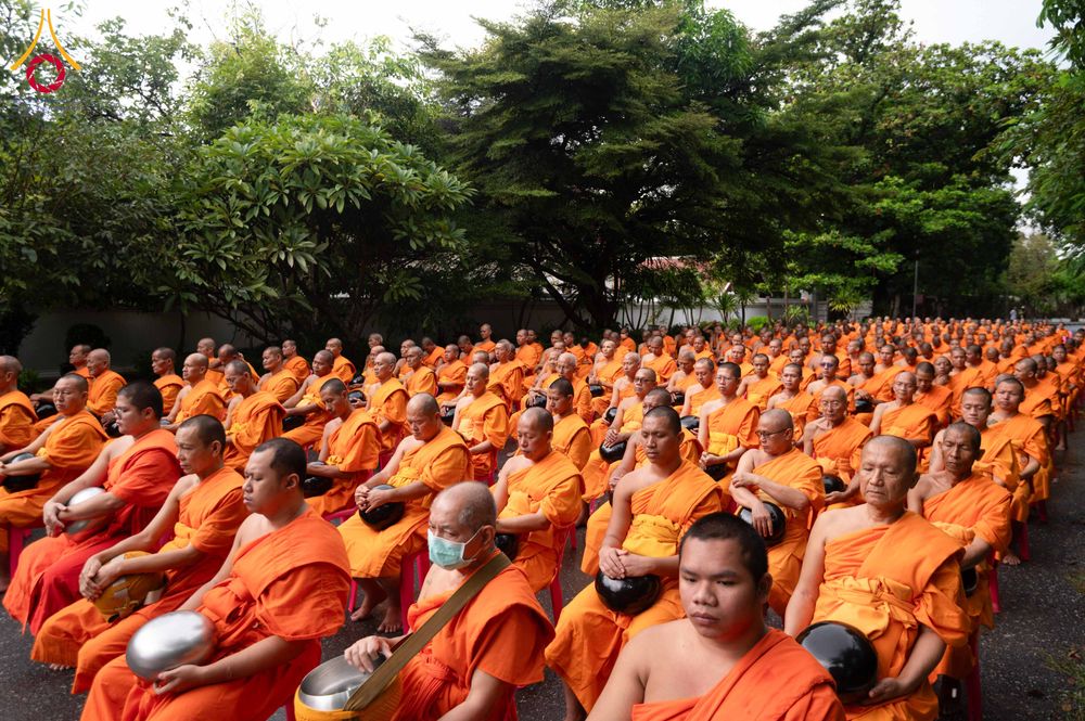 สามเณรจำนวนมากในจีวรสีส้มถือบาตรนั่งเรียงแถวในพื้นที่กลางแจ้ง