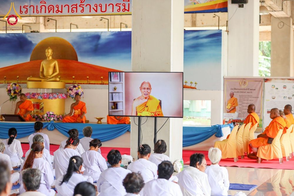 กลุ่มผู้เข้าร่วมอบรมในชุดสีขาวนั่งรับฟังธรรมะผ่านจอโทรทัศน์ภายในธุดงคสถาน