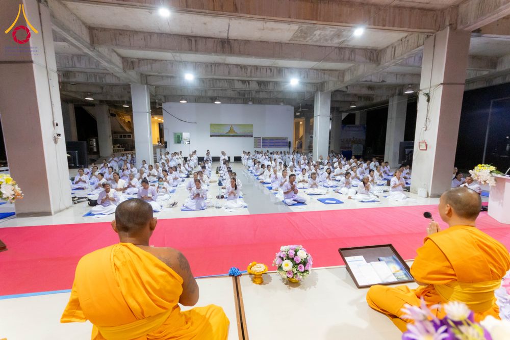 พระภิกษุสงฆ์นั่งหันหลังบนเวทีนำผู้เข้าร่วมโครงการสวมชุดขาวนั่งปฏิบัติธรรมภายในอาคาร
