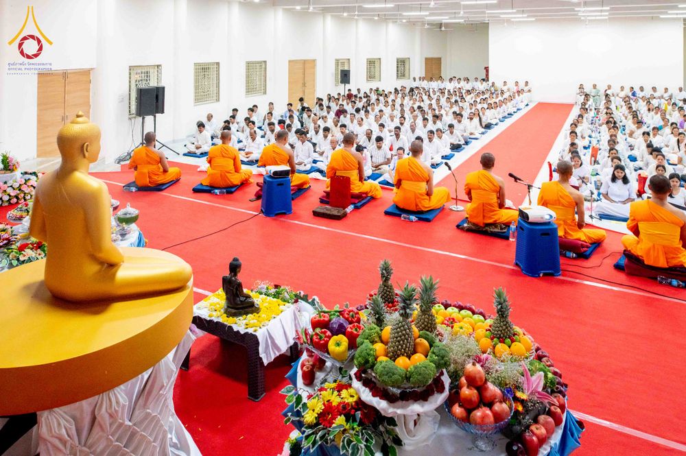 พระภิกษุและพุทธศาสนิกชนร่วมพิธีทางศาสนาในห้องโถงปูพรมสีแดง โดยมีพระพุทธรูปและเครื่องบูชาผลไม้อยู่ด้านหน้า