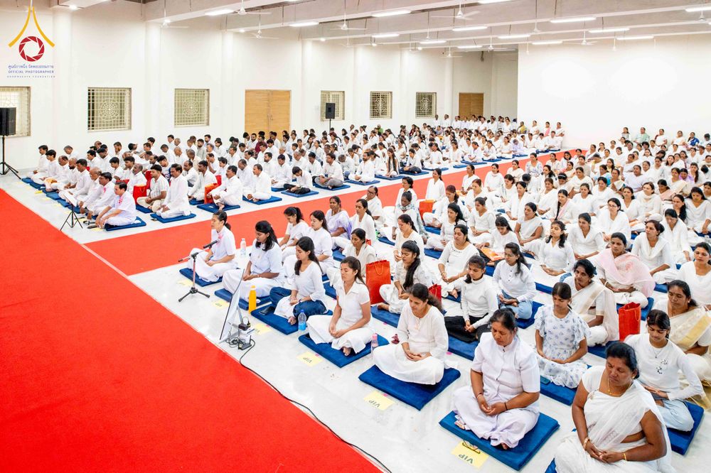 ผู้คนจำนวนมากสวมชุดขาวนั่งสมาธิอย่างสงบภายในห้องโถงขนาดใหญ่