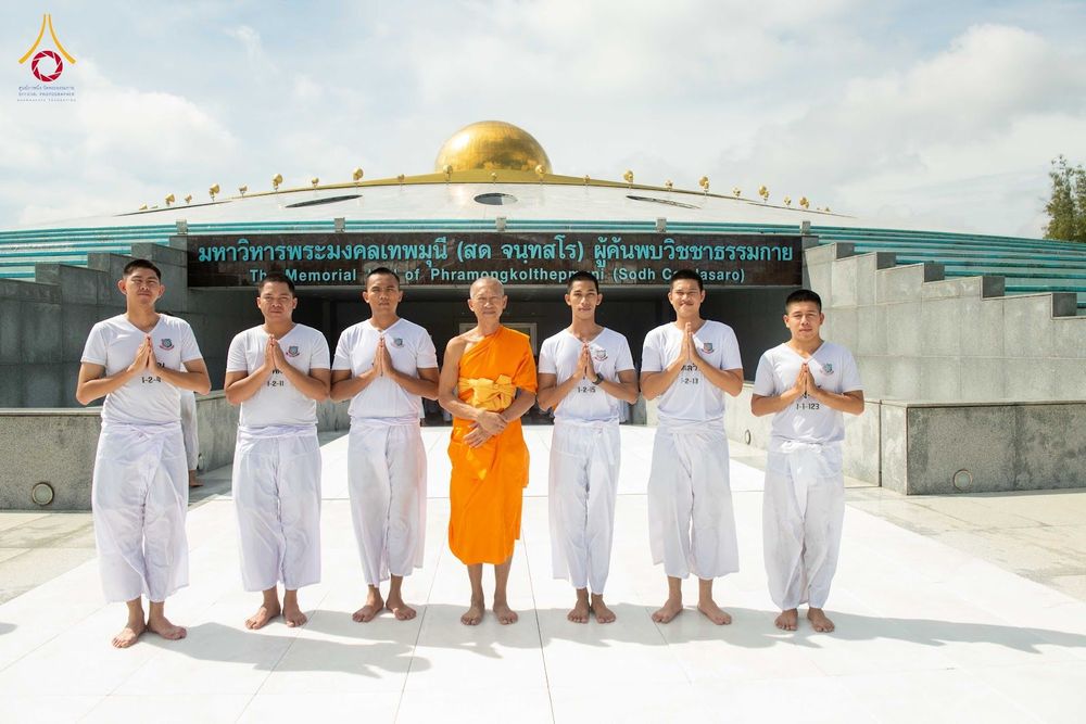 พระสงฆ์ยืนตรงกลางล้อมรอบด้วยนักเรียนนายสิบตำรวจในชุดขาวพนมมือไหว้หน้ามหาวิหารพระมงคลเทพมุนี