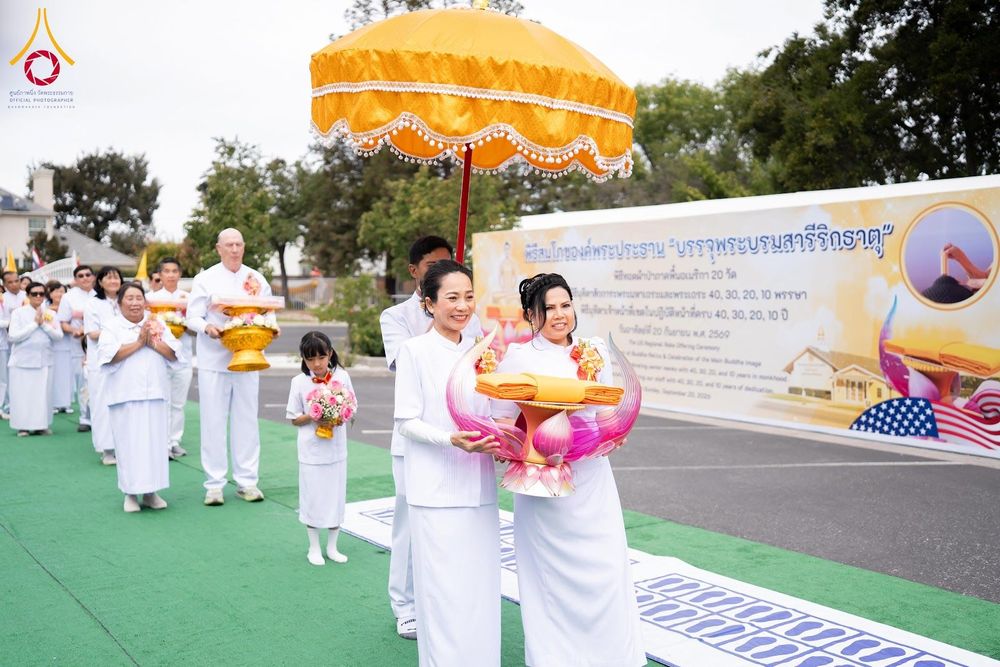 กลุ่มคนสวมชุดขาวถือพานพุ่มและผ้าไตรเดินขบวนในพิธีทางศาสนาพุทธกลางแจ้ง