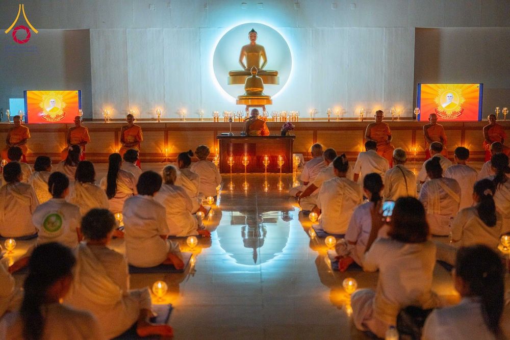กลุ่มคนชุดขาวนั่งสมาธิในห้องโถงที่มีพระพุทธรูปและแสงเทียนประดับ