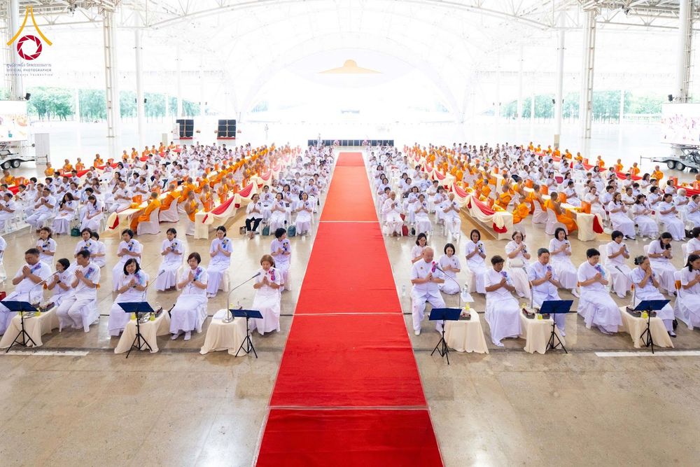 พุทธศาสนิกชนและพระภิกษุสงฆ์นั่งร่วมพิธีทางศาสนาภายในอาคารขนาดใหญ่ที่มีทางเดินปูพรมแดงตรงกลาง
