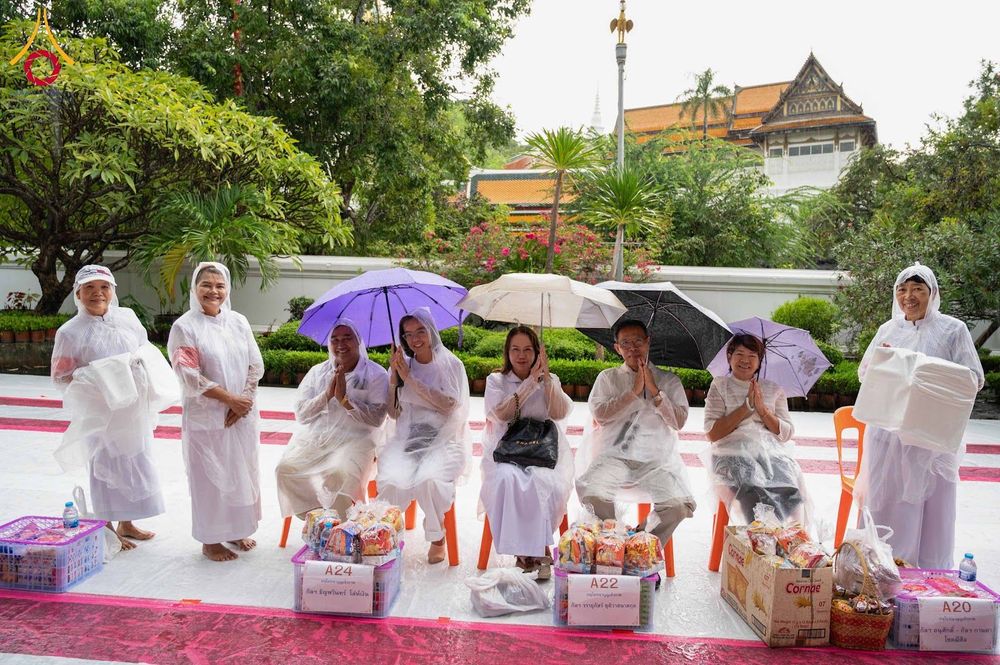 กลุ่มคนสวมชุดสีขาวและเสื้อกันฝนใส นั่งและยืนถือร่มพร้อมตะกร้าของตักบาตรวางอยู่ด้านหน้า