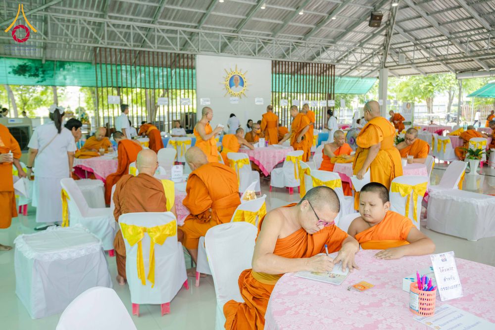พระภิกษุและผู้เข้าร่วมงานกำลังนั่งทำกิจกรรมสัมมนาภายในอาคารที่มีการจัดโต๊ะเก้าอี้คลุมผ้าสีชมพูและสีขาว