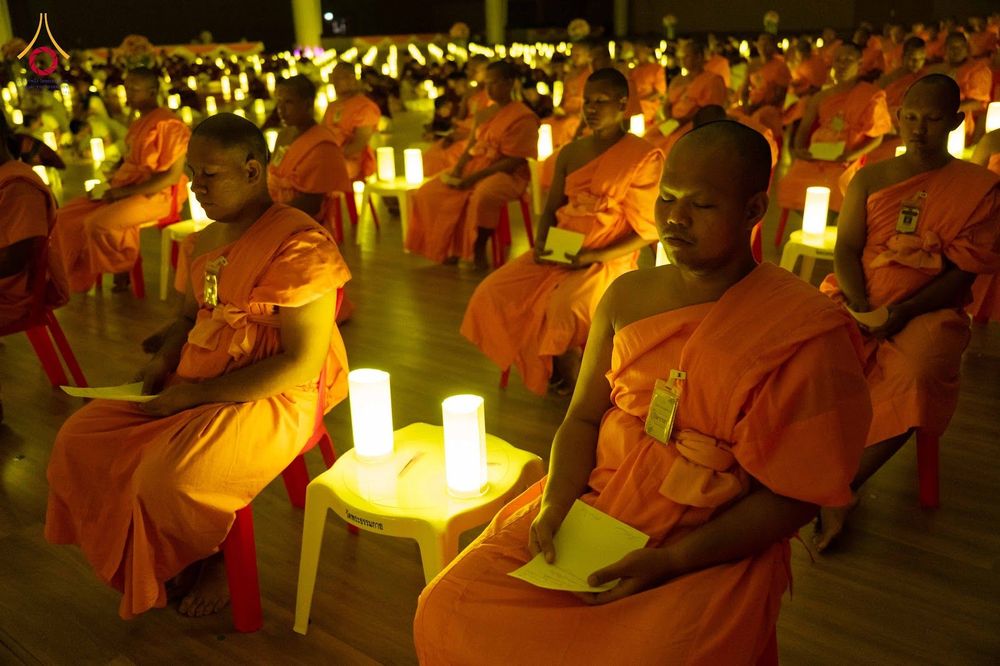 พระภิกษุสงฆ์หลายรูปนั่งสมาธิและถือกระดาษในมือท่ามกลางแสงเทียนภายในห้องโถง