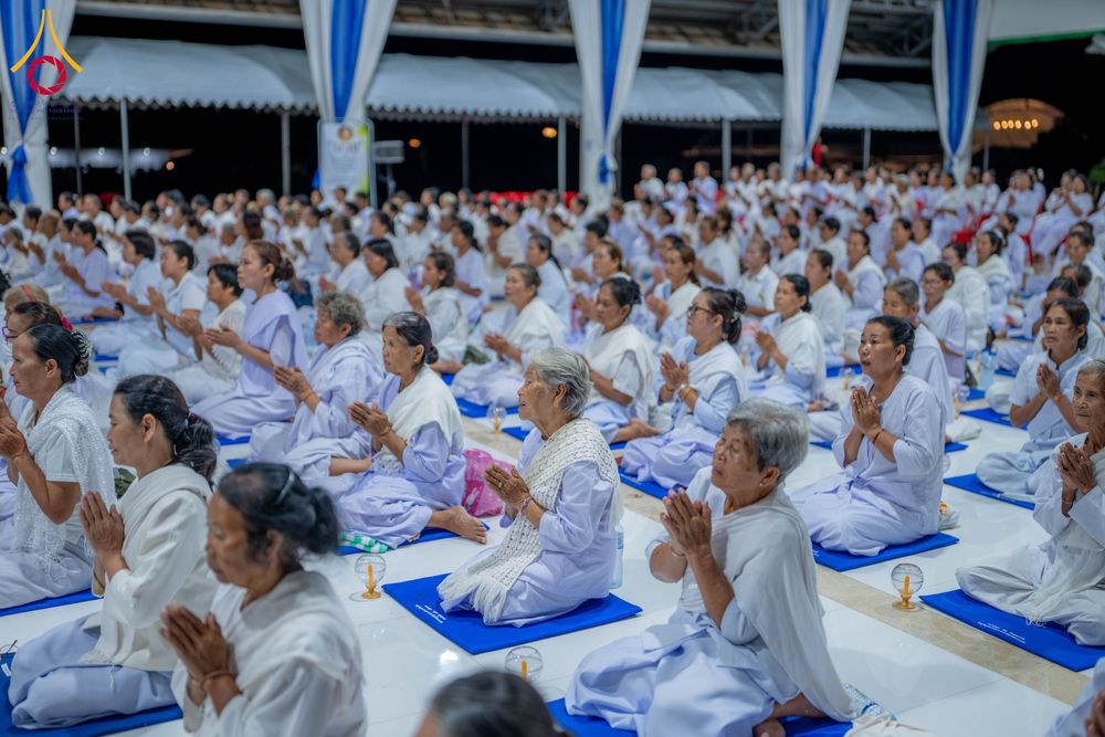 กลุ่มคนสวมชุดขาวนั่งพนมมือปฏิบัติธรรมบนเบาะรองนั่งสีน้ำเงินภายในอาคาร