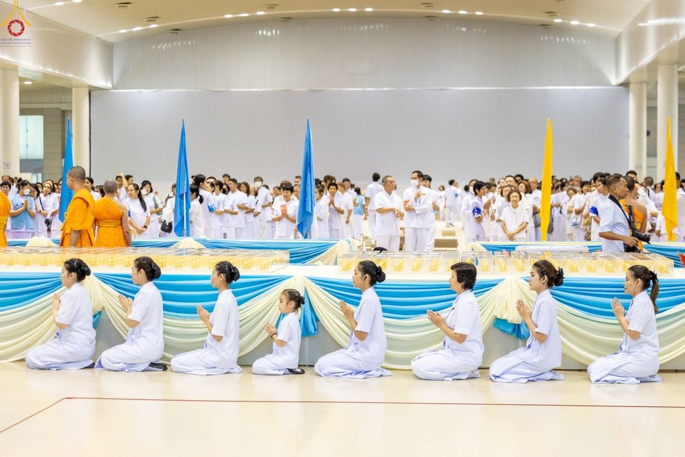 กลุ่มคนในชุดขาวนั่งพนมมือเรียงแถวหน้าโต๊ะวางพระของขวัญในพิธีทางศาสนา