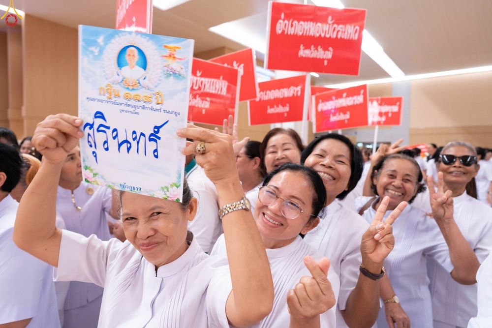กลุ่มคนสวมชุดสีขาวถือป้ายชื่อจังหวัดและป้ายงานบุญกฐินด้วยใบหน้ายิ้มแย้ม