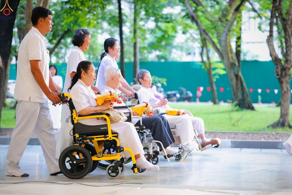 ผู้สูงอายุสวมชุดขาวนั่งบนรถเข็นมีเจ้าหน้าที่ช่วยเข็นในบริเวณวัด