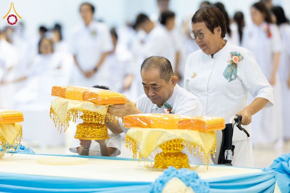พุทธศาสนิกชนในชุดขาวกำลังร่วมพิธีทอดผ้าป่า โดยมีชายและหญิงช่วยกันประคองพานพุ่มที่วางผ้าไตรสีเหลืองทอง