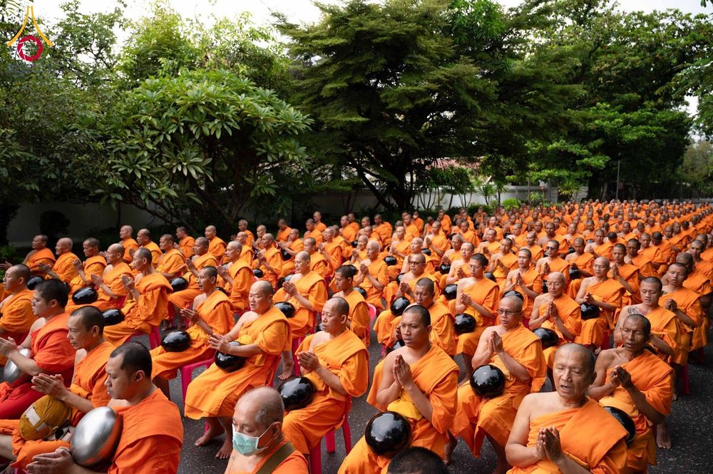 กลุ่มสามเณรจำนวนมากในจีวรสีส้มกำลังนั่งพนมมือเตรียมตัวในพิธีตักบาตรท่ามกลางบรรยากาศร่มรื่น