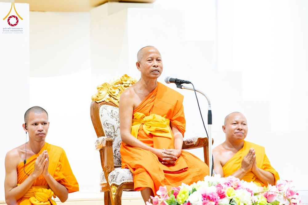 พระสงฆ์สามรูปในจีวรสีส้มกำลังประกอบพิธีทางศาสนา โดยรูปกลางนั่งบนเก้าอี้ไม้แกะสลักและกำลังพูดผ่านไมโครโฟน