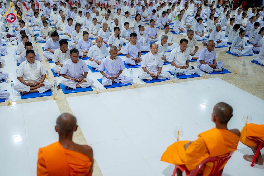 กลุ่มคนสวมชุดขาวนั่งสมาธิบนเบาะรองนั่งในอาคาร โดยมีพระภิกษุสองรูปนั่งอยู่ด้านหน้า