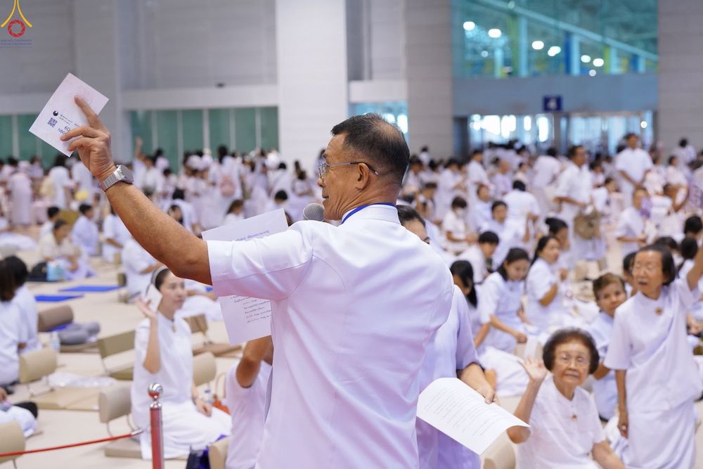 ชายสวมชุดขาวถือเอกสารและไมโครโฟนท่ามกลางผู้เข้าร่วมงานจำนวนมากในห้องโถง