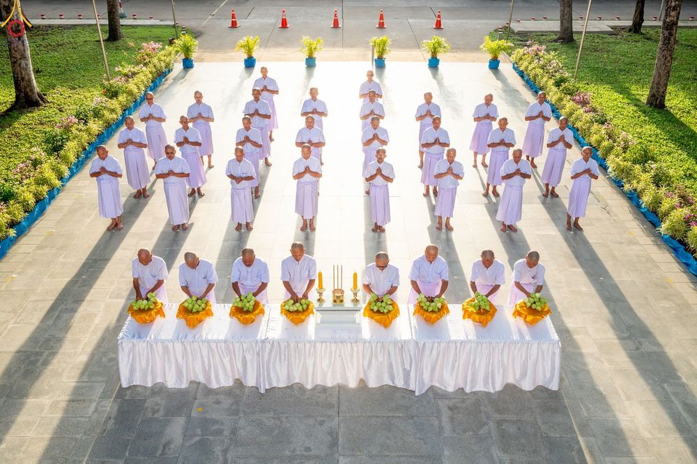 กลุ่มผู้เข้าพิธีบรรพชาอุปสมบทในชุดสีขาว ยืนเรียงแถวหน้าโต๊ะวางพานดอกไม้เพื่อประกอบพิธีกรรมทางศาสนา