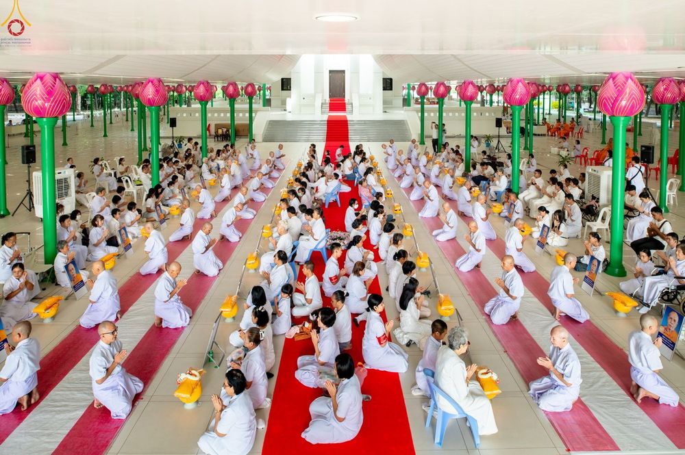 กลุ่มคนในชุดสีขาวนั่งพนมมือเรียงแถวภายในอาคารที่มีเสารูปดอกบัวสีชมพู เพื่อร่วมพิธีบรรพชาอุปสมบท