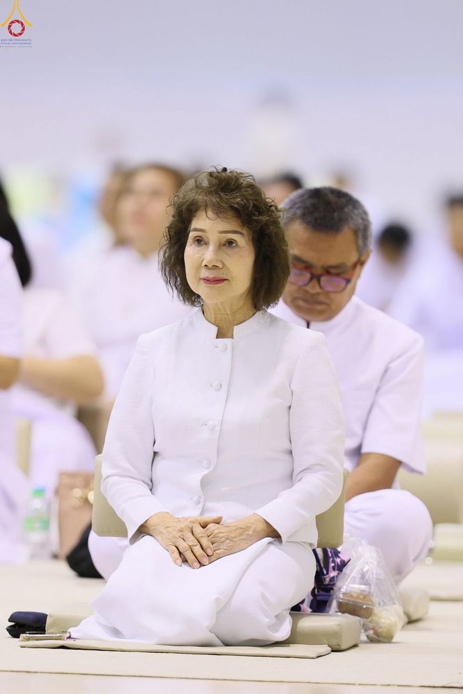 หญิงวัยกลางคนสวมชุดสีขาวนั่งพับเพียบร่วมกิจกรรมในห้องโถง