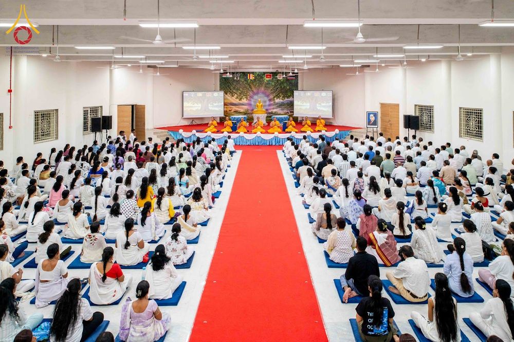 ผู้คนจำนวนมากสวมชุดขาวนั่งปฏิบัติธรรมในหอประชุมที่มีพรมแดงปูตรงกลางและพระสงฆ์นั่งบนเวทีด้านหน้า