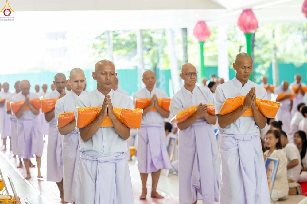 ผู้ชายหลายคนสวมชุดสีขาวถือผ้าไตรจีวรสีเหลืองพนมมือในพิธีทางศาสนา