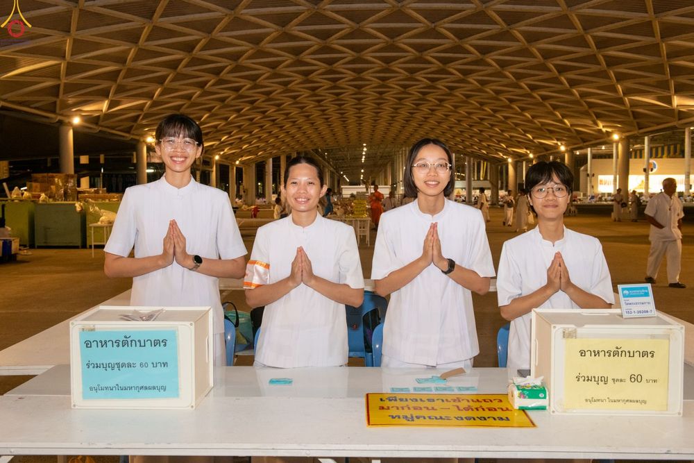 อาสาสมัครหญิงสี่คนสวมชุดสีขาวพนมมือยืนประจำจุดบริการอาหารตักบาตรภายในอาคารขนาดใหญ่