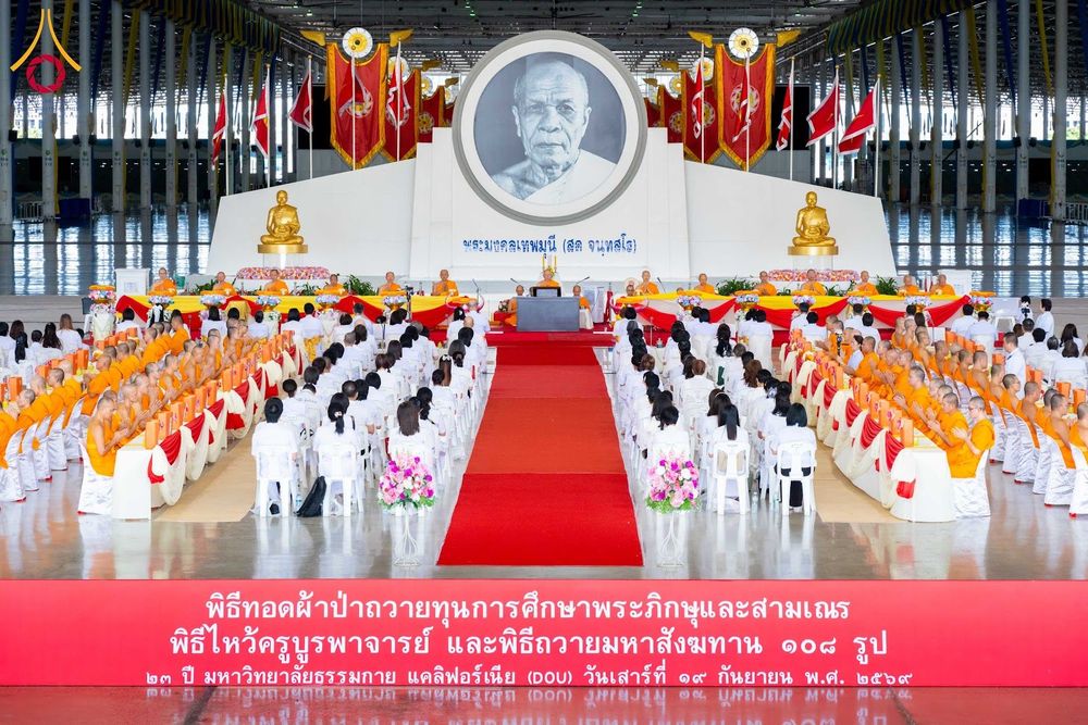 พุทธศาสนิกชนและพระภิกษุสงฆ์จำนวนมากเข้าร่วมพิธีทางศาสนาภายในสภาธรรมกายสากล โดยมีฉากหลังเป็นภาพพระมงคลเทพมุนีและธงประดับตกแต่งอย่างสวยงาม
