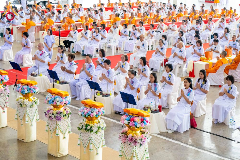 พุทธศาสนิกชนชุดขาวนั่งพนมมือร่วมพิธีทางศาสนาภายในสภาธรรมกายสากล โดยมีพานพุ่มดอกไม้ตั้งอยู่ด้านหน้า