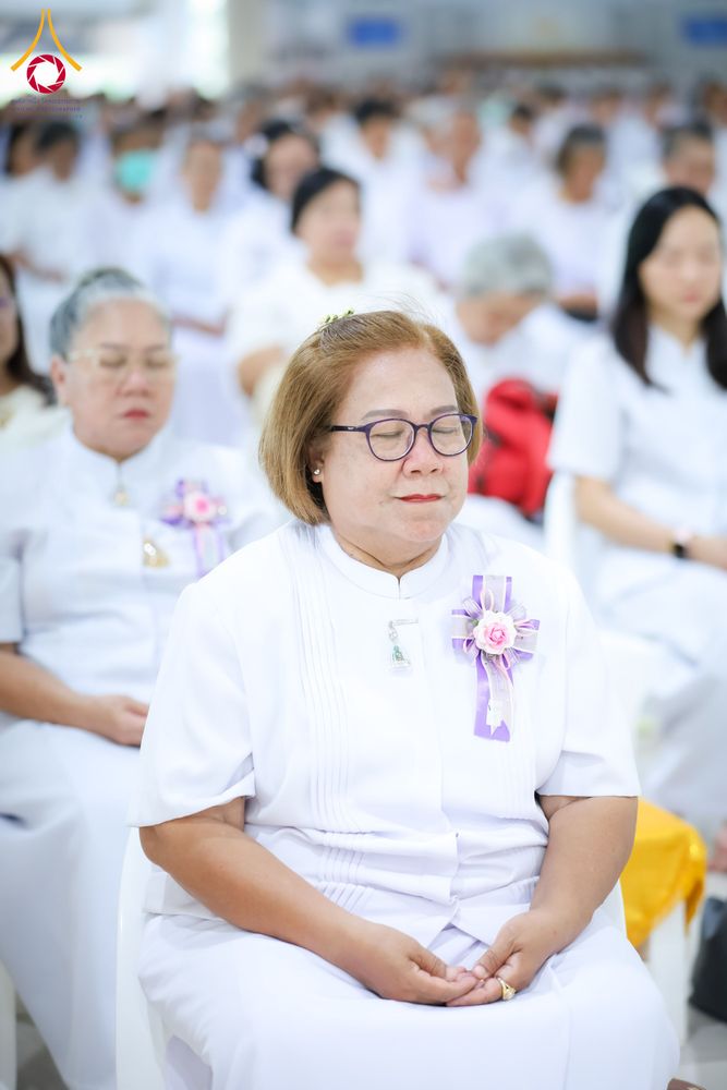 หญิงวัยกลางคนสวมชุดขาวและแว่นตานั่งหลับตาทำสมาธิร่วมกับกลุ่มคนในชุดขาวภายในอาคาร