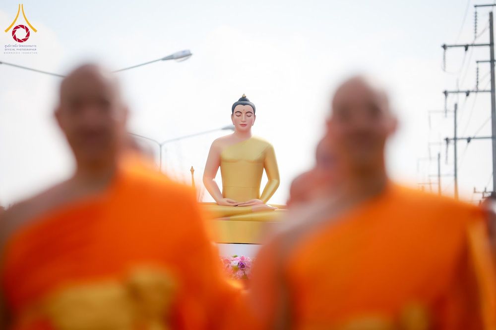 พระพุทธรูปสีทองปางสมาธิโดยมีพระภิกษุสงฆ์ในจีวรสีส้มเดินผ่านอยู่ด้านหน้าในลักษณะภาพเบลอ