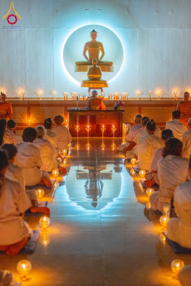 กลุ่มผู้เข้าร่วมโครงการสวมชุดขาวนั่งปฏิบัติธรรมภายในอาคารที่มีแสงเทียนวางเรียงรายบนพื้นและมีพระภิกษุนั่งเป็นประธานด้านหน้า