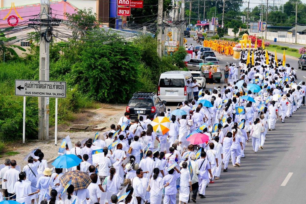 กลุ่มคนสวมชุดขาวจำนวนมากกำลังเดินขบวนไปตามถนนในพิธีทางศาสนา โดยมีขบวนพระภิกษุสงฆ์และรถบรรทุกองค์พระประธานนำหน้า