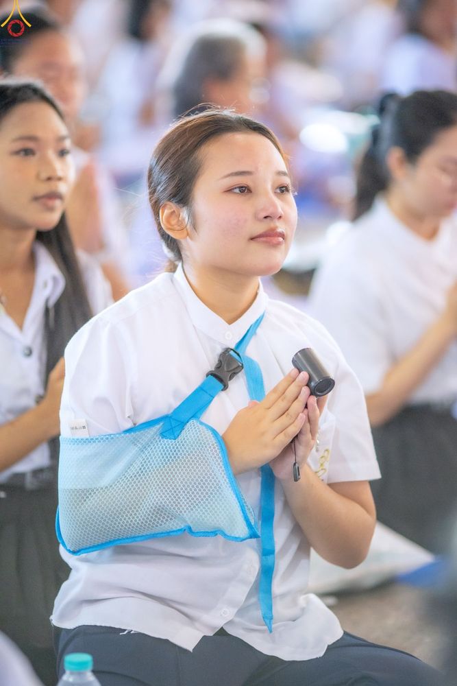 หญิงสาวสวมชุดสีขาวพนมมือและมีเฝือกอ่อนพยุงแขนขวาในพิธีการ