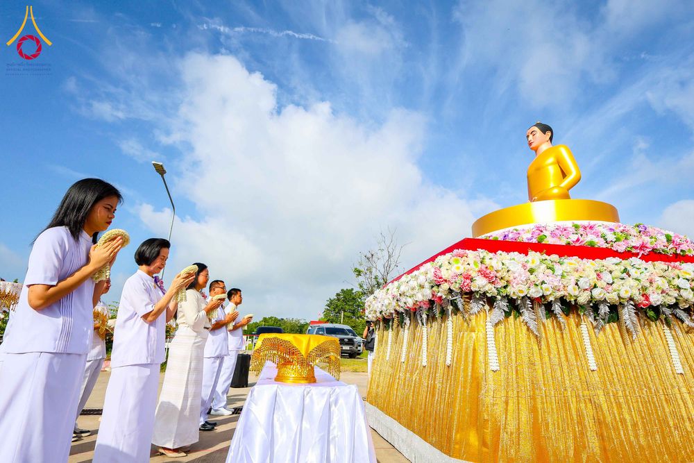 กลุ่มคนสวมชุดขาวกำลังไหว้พระพุทธรูปองค์สีทองที่ประดิษฐานบนฐานตกแต่งด้วยดอกไม้กลางแจ้ง