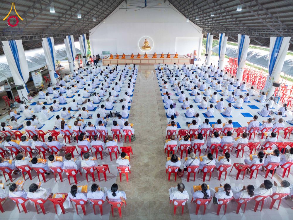 กลุ่มผู้เข้าร่วมโครงการสวมชุดขาวนั่งเรียงแถวอย่างเป็นระเบียบภายในอาคารปฏิบัติธรรมขนาดใหญ่ โดยมีพระภิกษุสงฆ์นั่งอยู่บนเวทีด้านหน้า