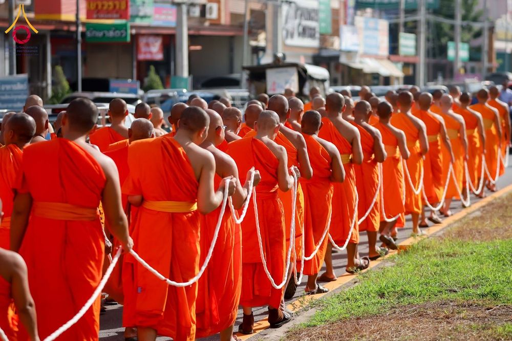 พระภิกษุสงฆ์จำนวนมากสวมจีวรสีส้มเดินเรียงแถวถือสายสิญจน์บนถนน