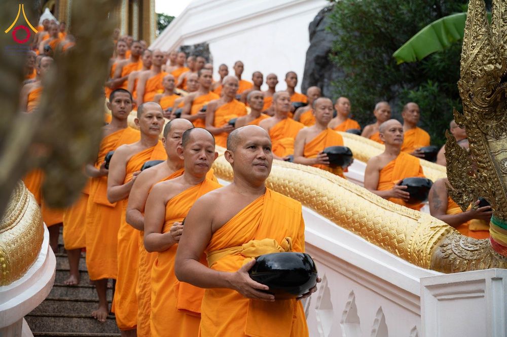 กลุ่มพระภิกษุและสามเณรห่มจีวรสีส้มถือบาตรเดินเรียงแถวลงจากบันไดวัดที่มีราวบันไดรูปพญานาค