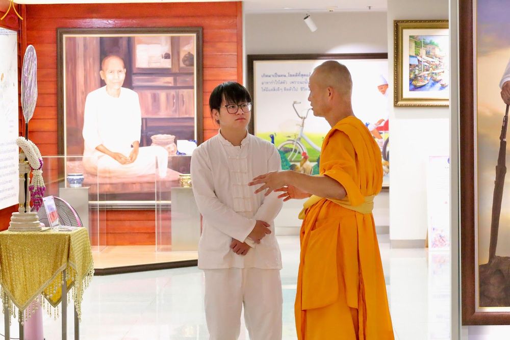 พระภิกษุสนทนากับชายชาวต่างชาติในชุดสีขาวภายในห้องจัดแสดงภาพ