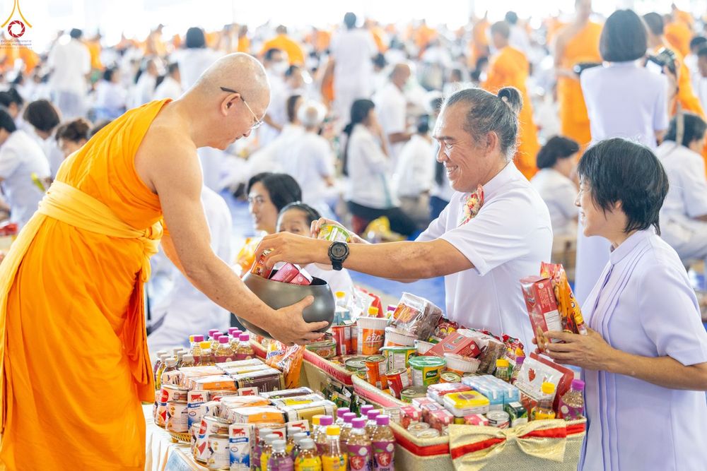 ชายและหญิงสวมชุดสีขาวกำลังตักบาตรอาหารแห้งใส่บาตรพระภิกษุสงฆ์ท่ามกลางพุทธศาสนิกชนจำนวนมาก