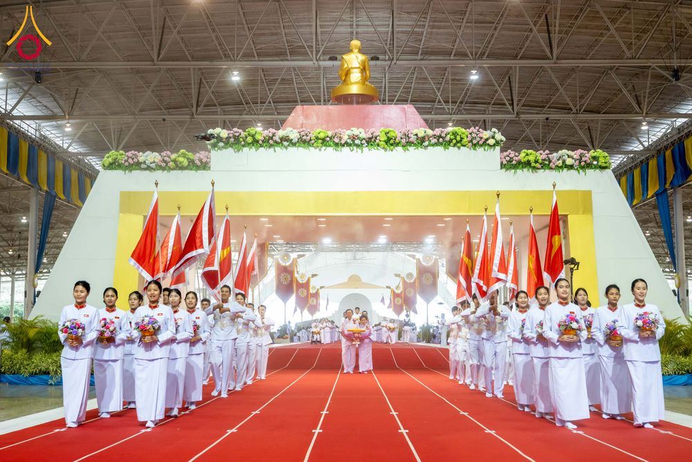 กลุ่มคนในชุดขาวถือธงและพานดอกไม้เดินแถวบนพรมแดงในอาคารขนาดใหญ่