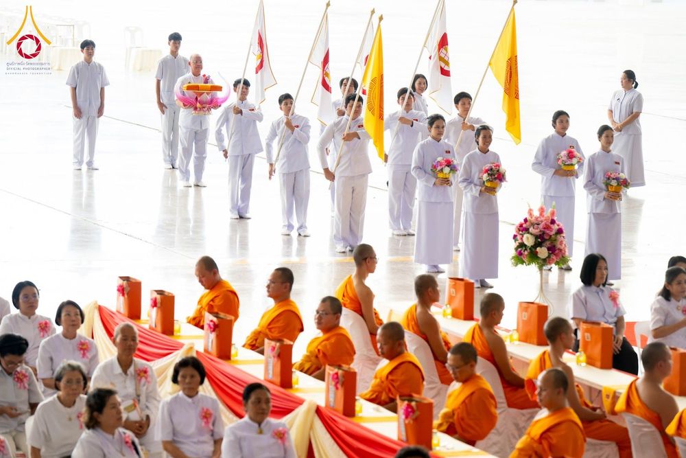ขบวนผู้ถือธงและพานดอกไม้เดินนำหน้าพระภิกษุสงฆ์และพุทธศาสนิกชนชุดขาวในพิธีทางศาสนา
