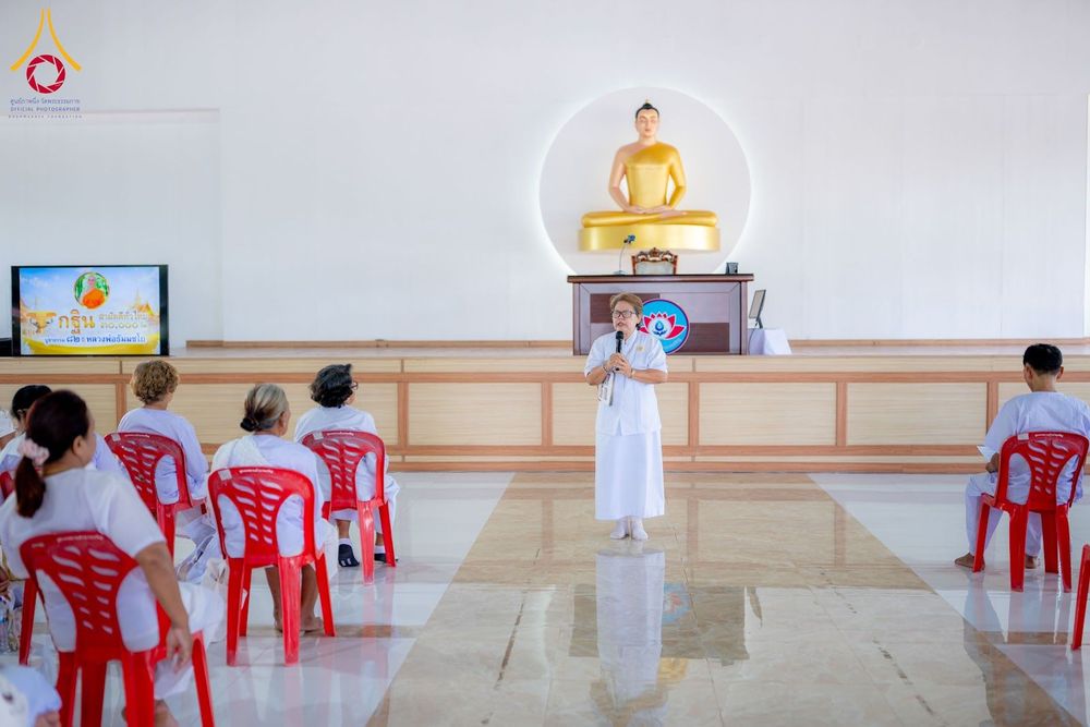 วิทยากรหญิงสวมชุดขาวกำลังบรรยายธรรมให้ผู้เข้าร่วมโครงการอบรมอุบาสิกาแก้วที่นั่งบนเก้าอี้พลาสติกสีแดงภายในห้องโถง