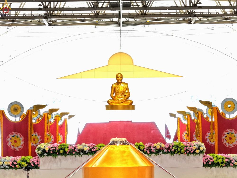 รูปหล่อทองคำของพระภิกษุในท่านั่งสมาธิบนรัตนบัลลังก์ มีฉากหลังเป็นธงสีแดงและดอกไม้ประดับ